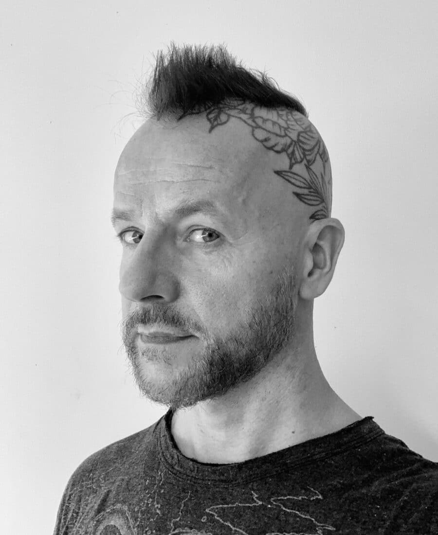 A person with a side-shaved head featuring tattoos and a short, spiked mohawk hairstyle, wearing a dark shirt, looking sideways against a plain background.