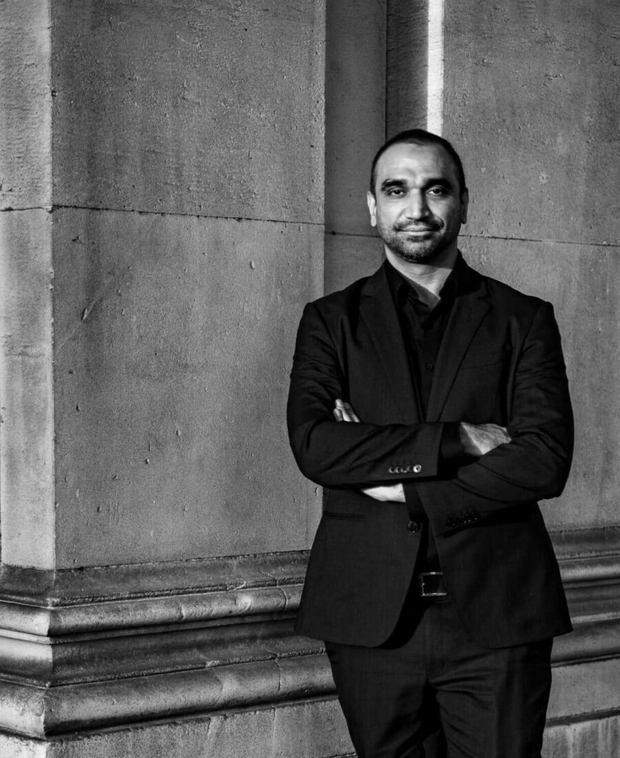 Black and white image of a person in a dark suit standing against a stone wall with arms crossed, looking at the camera.