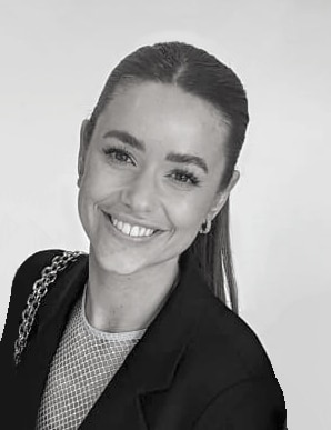Smiling person with long hair in a ponytail, wearing a dark blazer over a mesh top, against a plain background.
