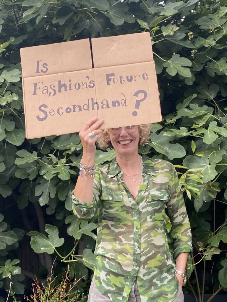 A person in a green camouflage shirt holds a cardboard sign reading, "Is Fashion's Future Secondhand?" while standing in front of leafy green plants.
