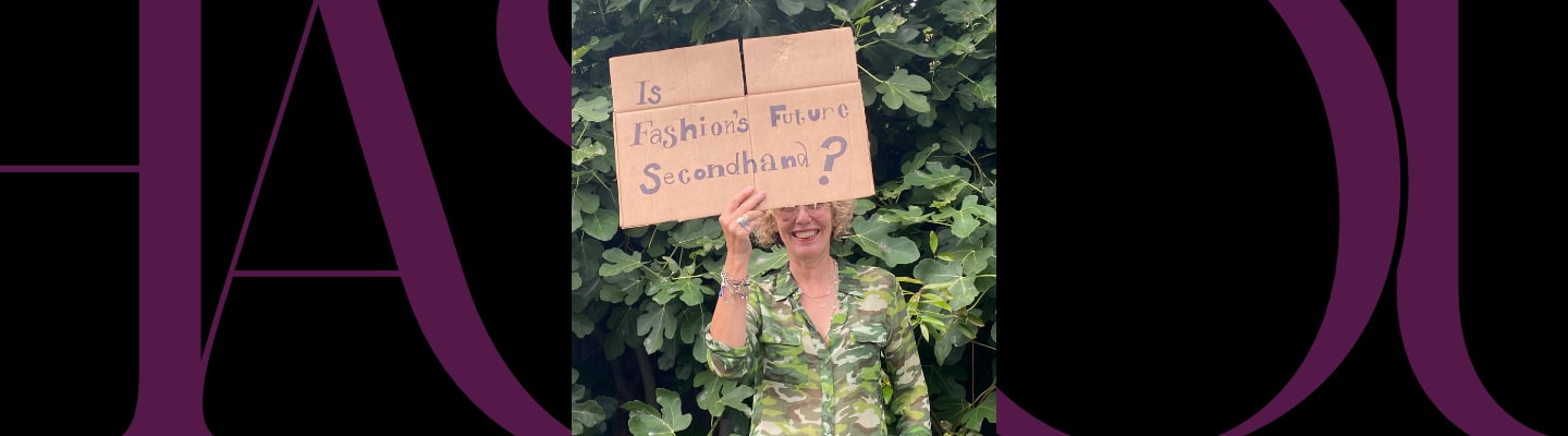 Person standing in front of leafy bushes holds a cardboard sign reading, "Is Fashion's Future Secondhand?.