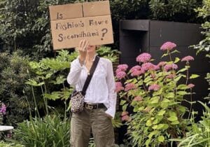 A person stands in a garden holding a cardboard sign reading “Is Fashion’s Future Secondhand?” while wearing a white shirt, cargo pants, yellow shoes, and a crossbody bag.