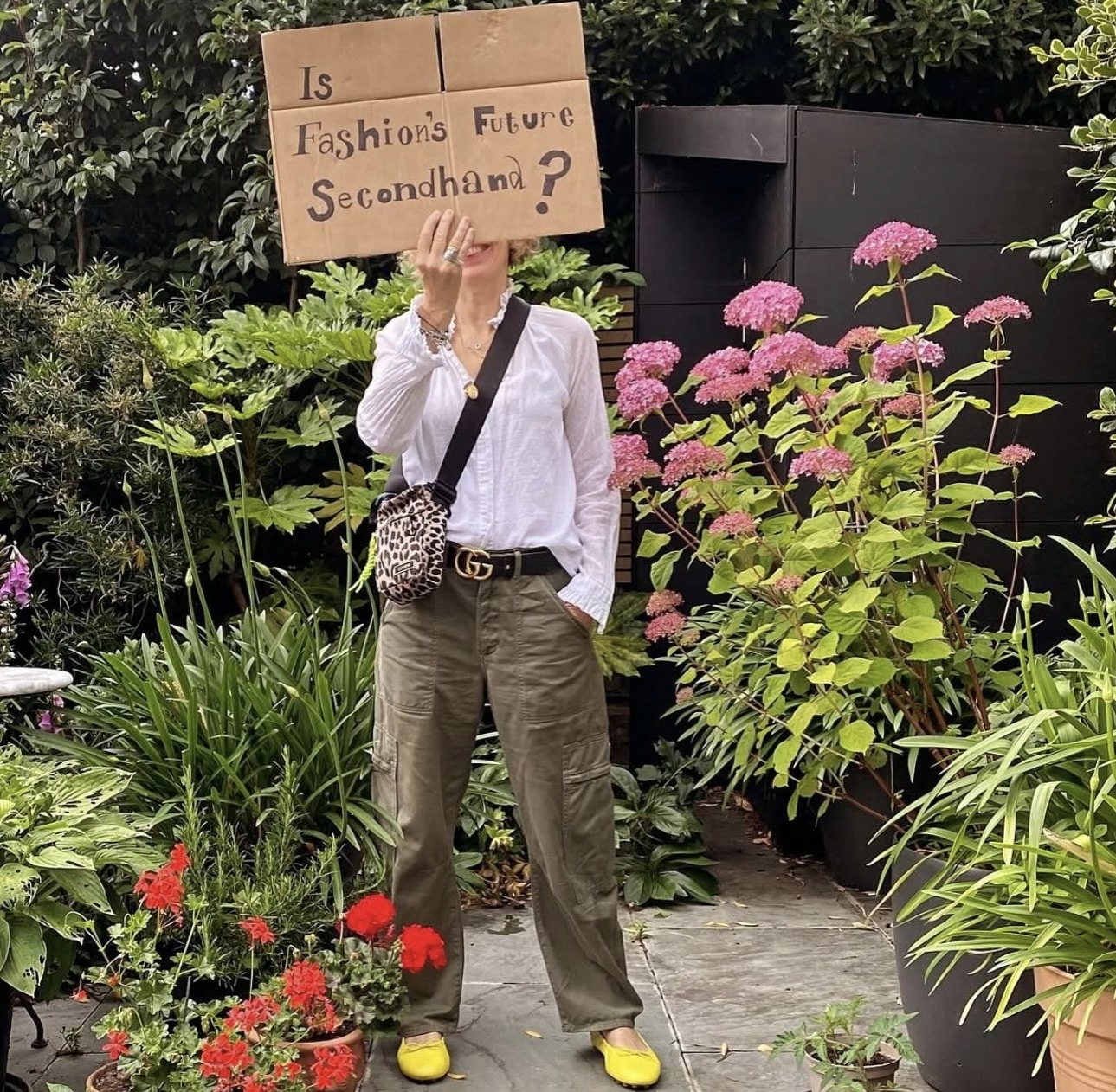 A person stands in a garden holding a cardboard sign reading “Is Fashion’s Future Secondhand?” while wearing a white shirt, cargo pants, yellow shoes, and a crossbody bag.