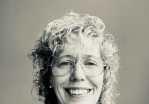 A person with curly hair and glasses smiles at the camera, wearing a leopard print shirt, against a plain background.