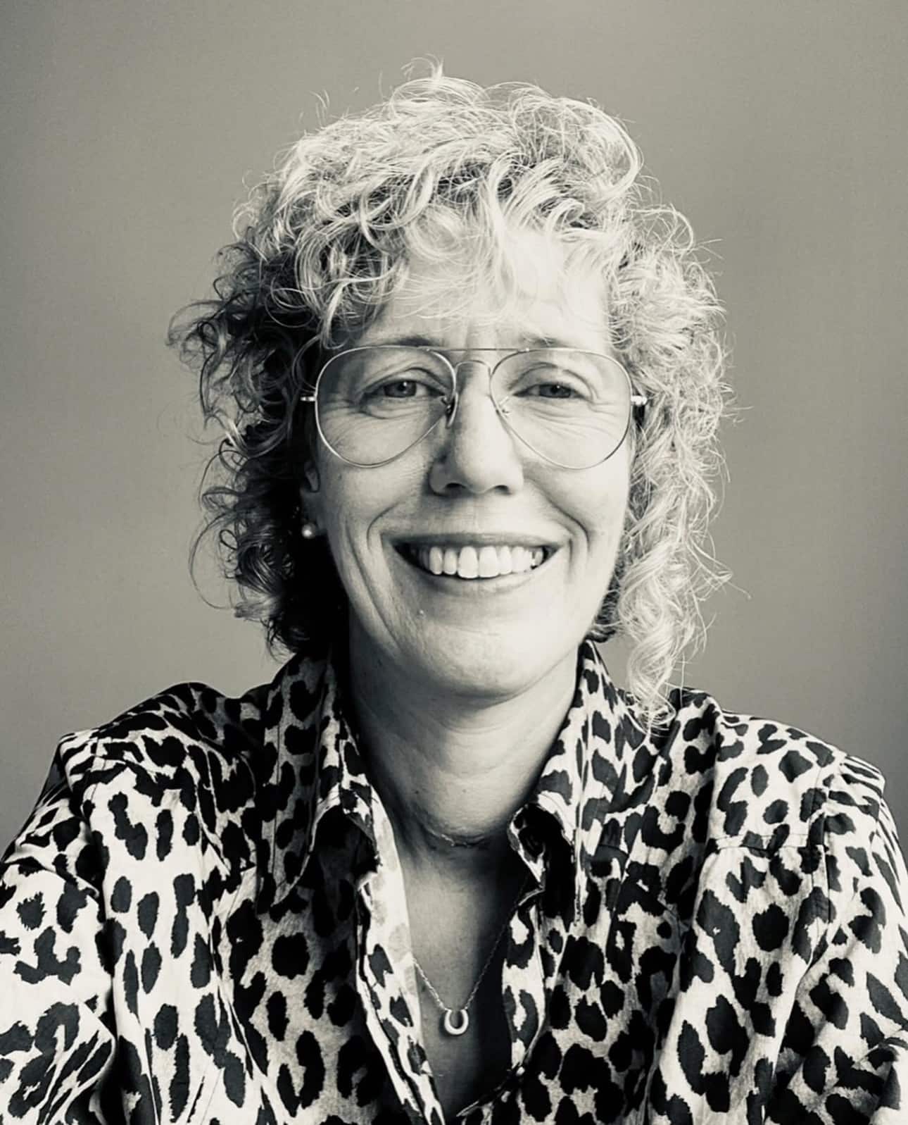 A person with curly hair and glasses smiles at the camera, wearing a leopard print shirt, against a plain background.