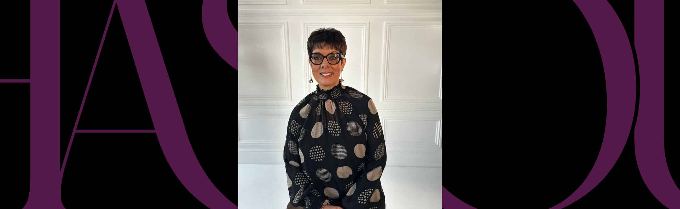 Person with short hair and glasses, wearing a patterned black top, sits smiling in front of a white paneled wall.