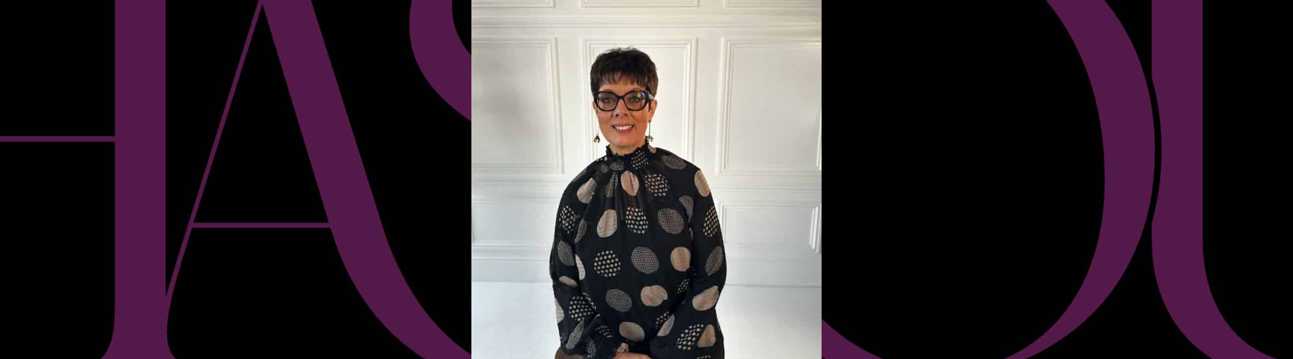 Person with short hair and glasses, wearing a patterned black top, sits smiling in front of a white paneled wall.