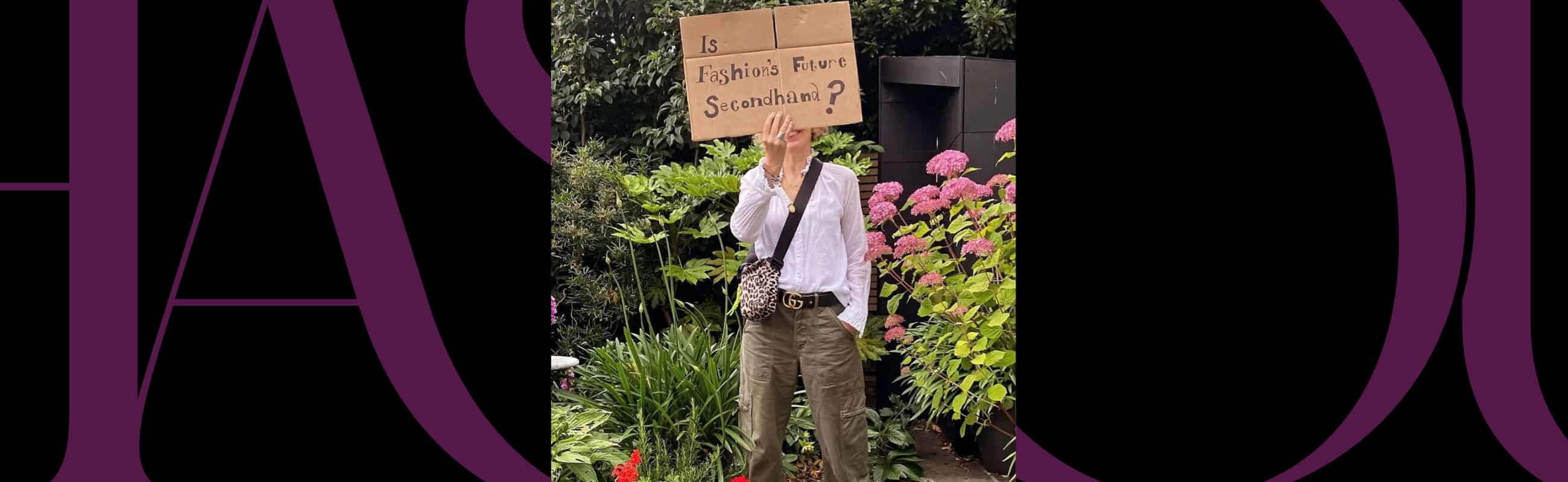 A person stands in a garden holding a cardboard sign that reads, "Is Fashion's Future Sustainable?" Pink flowers and greenery are visible in the background.
