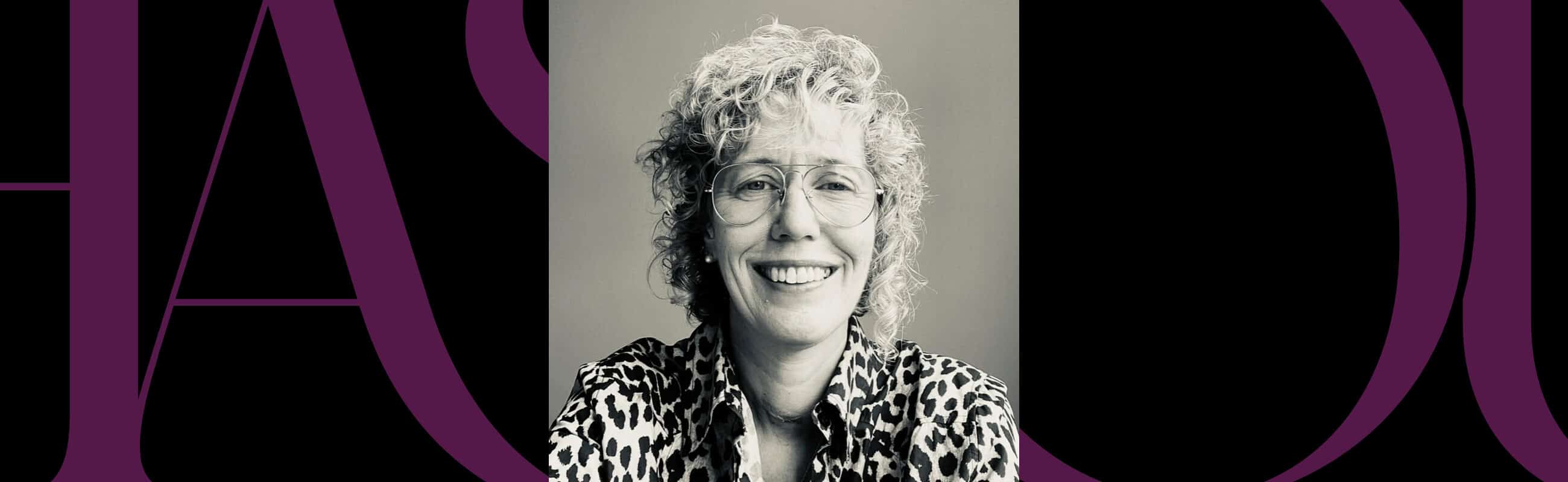 Black and white portrait of a smiling person with curly hair and glasses, wearing a leopard print shirt, centered on a background with large purple letters.