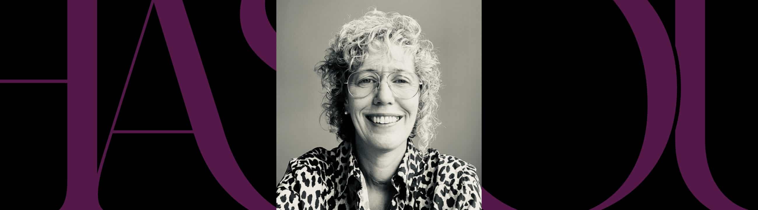 Black and white portrait of a smiling person with curly hair and glasses, wearing a leopard print shirt, centered on a background with large purple letters.
