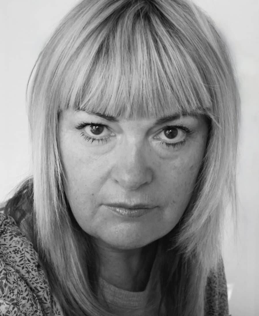 A black and white portrait of a woman with straight, light-colored hair and bangs, looking directly at the camera with a neutral expression.