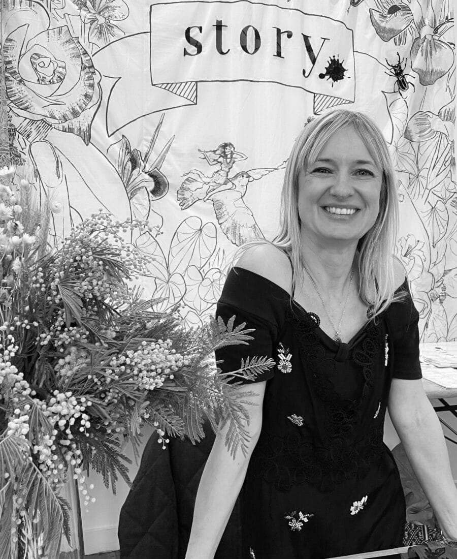 A woman stands behind a table, smiling, in front of a decorative backdrop that reads "story" with floral illustrations.