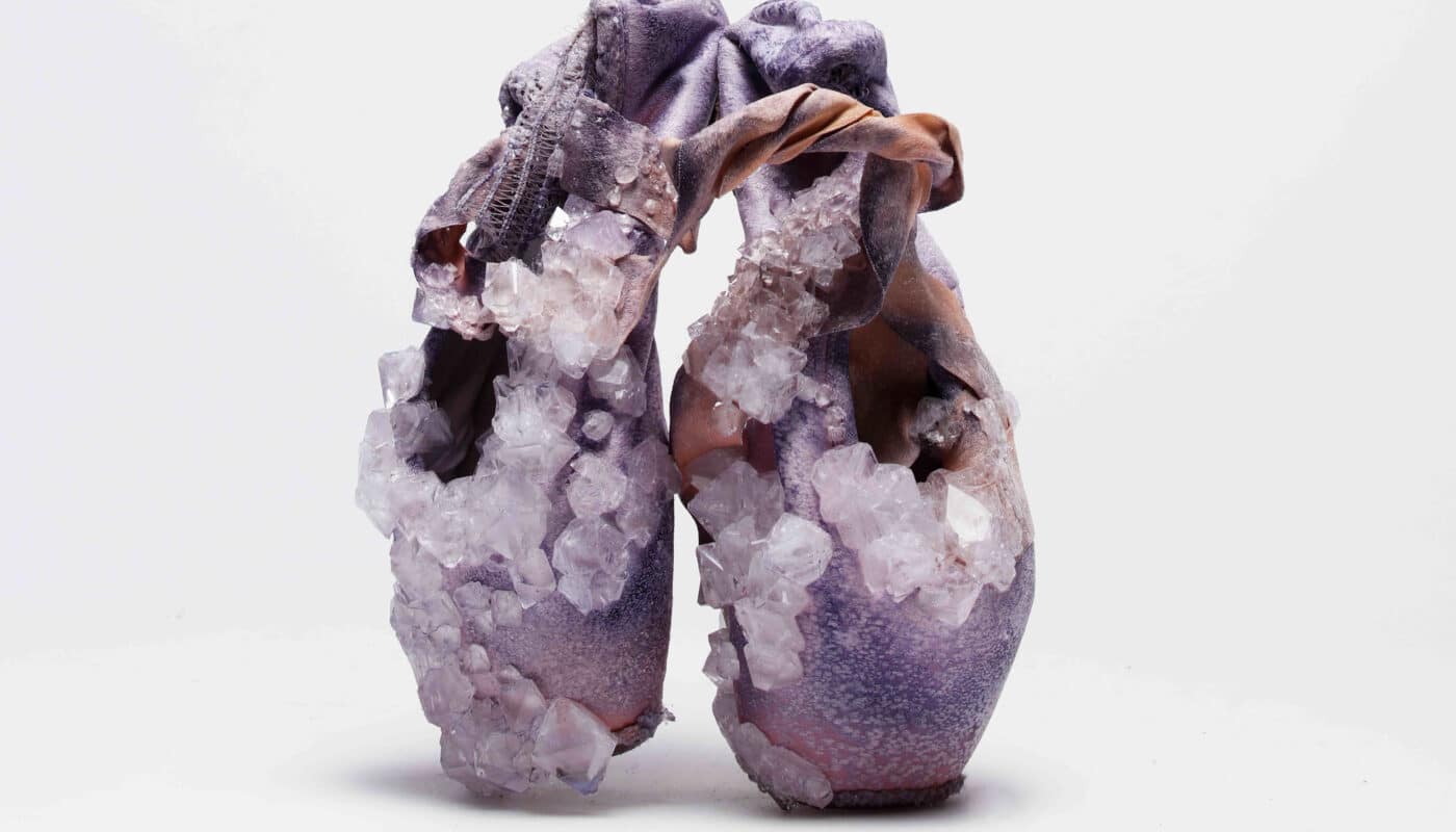 A pair of worn purple ballet pointe shoes encrusted with large, translucent crystals against a plain white background.