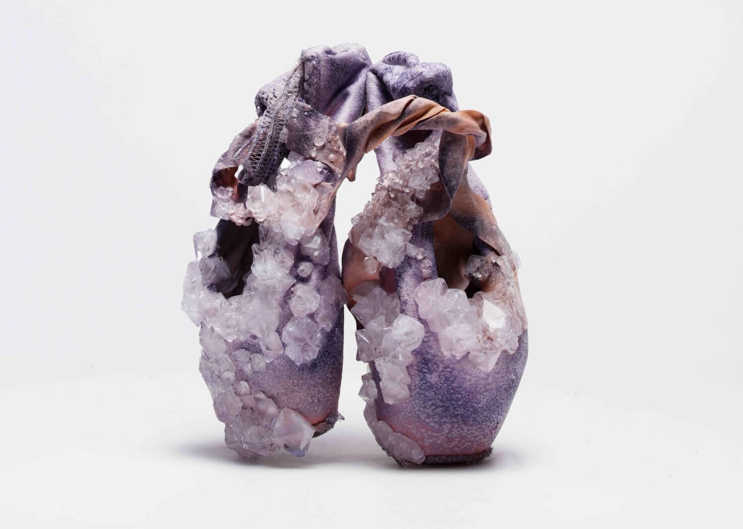 A pair of worn purple ballet pointe shoes encrusted with large, translucent crystals against a plain white background.
