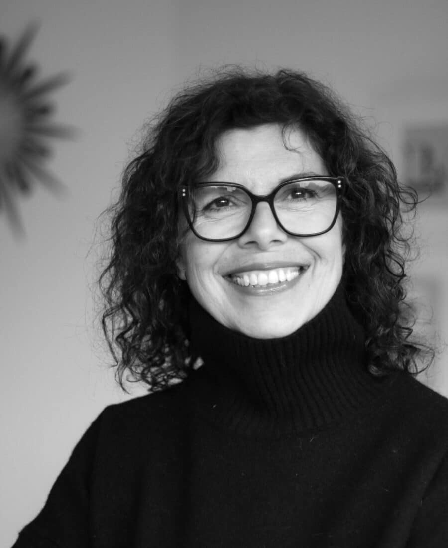 A woman with curly hair and glasses smiles at the camera, wearing a dark turtleneck sweater. The background is softly blurred.