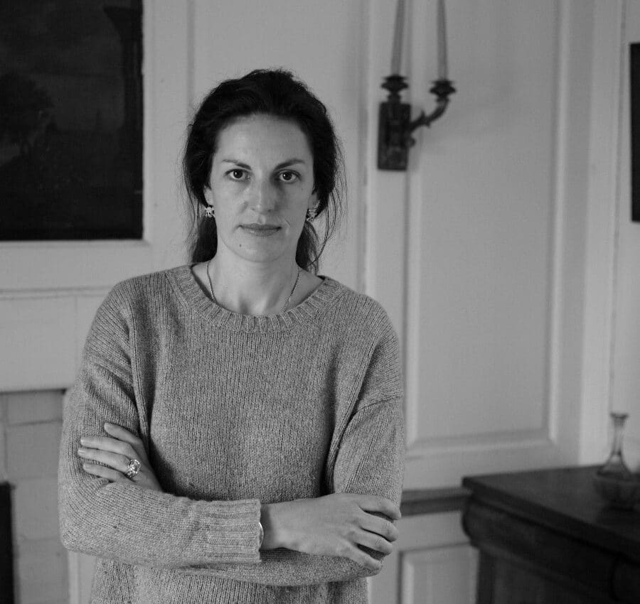 A woman with long dark hair and a jumper stands indoors with her arms crossed, looking at the camera. The room has framed art, a mantelpiece, and decorative items.